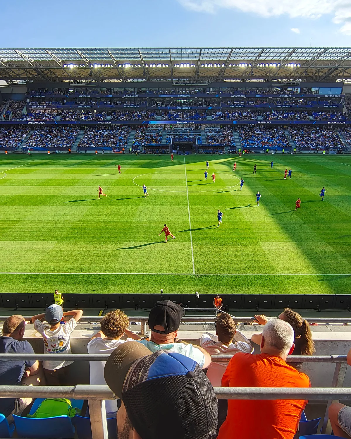 Vue depuis la tribune d’un stade plein : les deux équipes à l’échauffement sur la pelouse, des supporters au premier rang